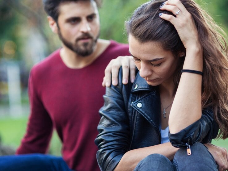 Ein Mann legt tröstend seine Hand auf die Schulter einer nachdenklich und traurig wirkenden Frau. Beide sitzen im Freien, die Frau hält ihren Kopf mit einer Hand und wirkt besorgt, während der Mann mitfühlend neben ihr sitzt.