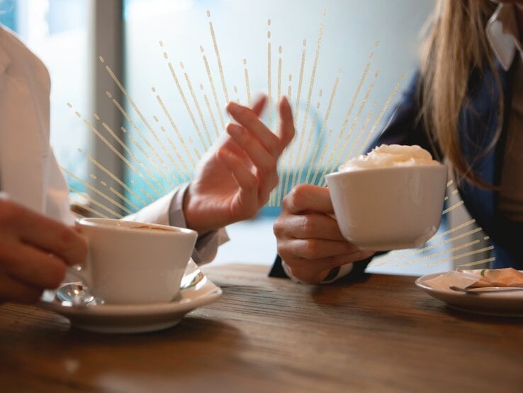 Zwei Personen sitzen in einem Café und halten Tassen mit Kaffee in den Händen, während sie sich unterhalten. Ihre Gesten und die Strahlen, die das Bild zieren, vermitteln eine energetische und positive Stimmung.