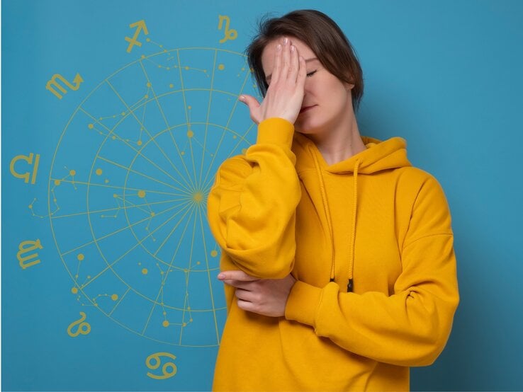 Eine Frau in einem gelben Hoodie steht vor einem blauen Hintergrund mit astrologischen Symbolen und hält sich frustriert die Hand vor das Gesicht. Ihre Haltung deutet auf Enttäuschung oder Erschöpfung hin, während das astrologische Rad im Hintergrund eine Verbindung zu Horoskopen oder astrologischen Themen andeutet.