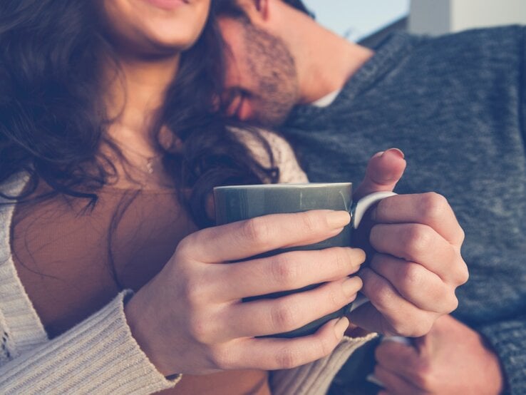 Ein Paar sitzt nah beieinander, während die Frau eine Tasse in beiden Händen hält. Der Mann lehnt seinen Kopf sanft an ihre Schulter, und beide wirken entspannt und verbunden. Die Frau lächelt leicht, während sie die Wärme der Tasse genießt. Das Bild vermittelt eine gemütliche, intime Atmosphäre und die Vertrautheit zwischen den beiden.