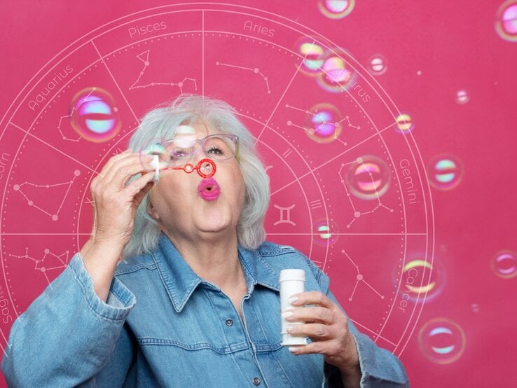 Eine ältere Frau mit weißem Haar und einer Brille bläst Seifenblasen in die Luft. Sie trägt eine Jeansjacke und steht vor einem pinkfarbenen Hintergrund, auf dem ein astrologisches Rad mit den Tierkreiszeichen angedeutet ist. Die Seifenblasen schweben vor ihr und reflektieren bunte Lichtpunkte, die eine spielerische und leichte Atmosphäre erzeugen.