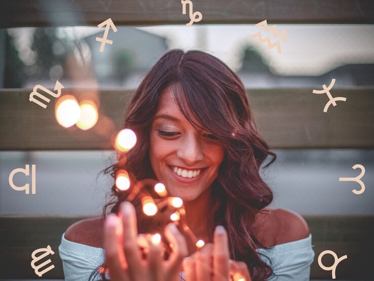 Eine lächelnde Frau mit dunklen Haaren hält leuchtende Lichter in den Händen, während um sie herum astrologische Symbole der Tierkreiszeichen schweben. Sie wirkt glücklich und entspannt, was eine positive und magische Atmosphäre vermittelt.