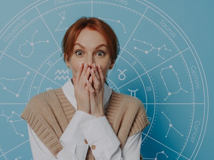 Eine überraschte Frau mit rotblonden Haaren hält sich die Hände vor den Mund und schaut mit großen Augen. Hinter ihr ist ein blauer Hintergrund mit einem astrologischen Rad und den Symbolen der Tierkreiszeichen zu sehen.
