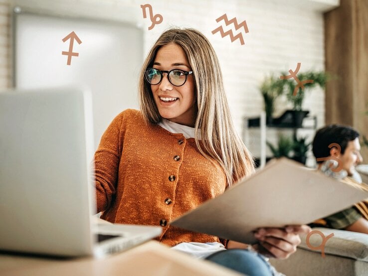 Eine Frau mit Brille und einem orangefarbenen Pullover sitzt an einem Laptop und hält einen Ordner in der Hand. Sie lächelt und wirkt konzentriert, während um sie herum astrologische Symbole schweben. Die Szene vermittelt eine produktive und motivierte Arbeitsatmosphäre mit einem Hauch von astrologischem Einfluss.