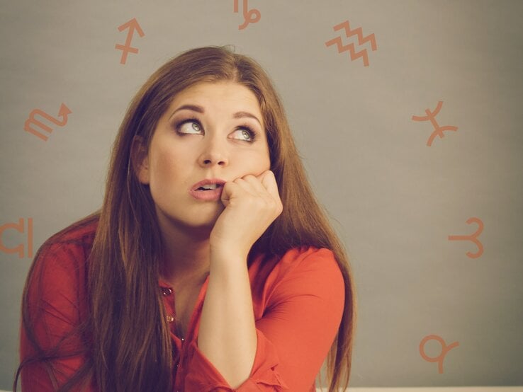 Eine nachdenkliche Frau in einem roten Oberteil stützt ihren Kopf auf die Hand und blickt skeptisch nach oben. Der Hintergrund ist neutral gehalten, während astrologische Symbole in warmen Tönen dezent um sie herum verteilt sind. Das Bild vermittelt Unsicherheit und Nachdenklichkeit, kombiniert mit einem Bezug zur Astrologie.