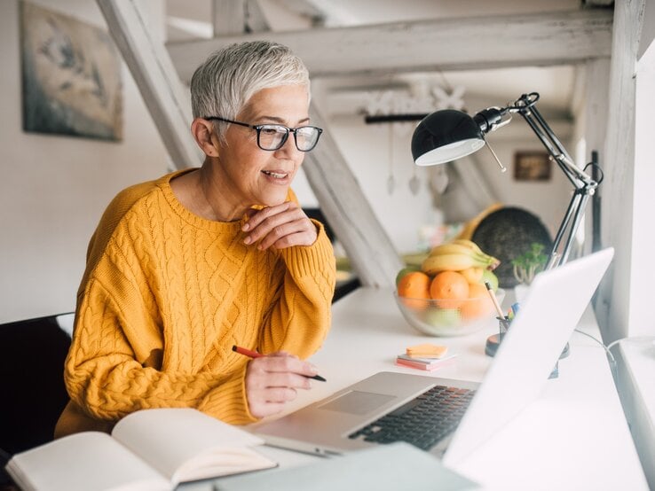 Eine ältere Frau mit grauem, kurzem Haar und Brille sitzt an einem Schreibtisch und arbeitet konzentriert an ihrem Laptop. Sie trägt einen gemütlichen gelben Strickpullover und hält einen Stift in der Hand. Auf dem Schreibtisch liegen ein aufgeschlagenes Notizbuch, Post-its und eine Schreibtischlampe. Im Hintergrund stehen eine Schale mit frischem Obst und dezente Einrichtungsdetails, die eine helle und freundliche Arbeitsatmosphäre schaffen.