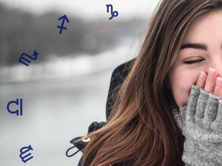 Eine junge Frau mit langen braunen Haaren, die eine dunkle Jacke und graue Wollhandschuhe trägt. Sie hält lächelnd ihre Hände an ihr Gesicht, während sie die Augen geschlossen hat. Im Hintergrund ist eine winterliche Landschaft mit verschwommenem Wasser und grauem Himmel zu sehen. Um sie herum schweben Symbole der Sternzeichen, wie Schütze und Skorpion.