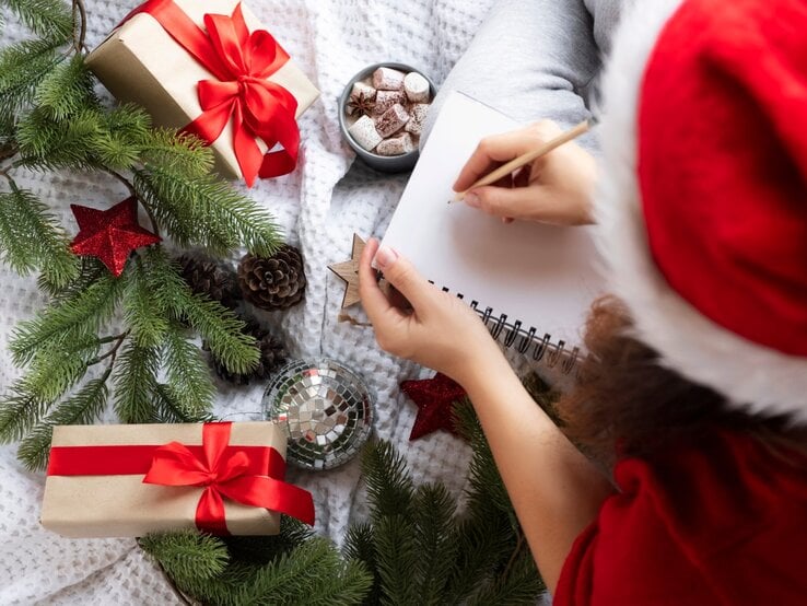 Ein weihnachtliches Bild zeigt eine Person, die in ein leeres Notizbuch schreibt. Sie trägt eine rote Weihnachtsmütze und sitzt auf einer kuscheligen, weißen Decke. Um die Person herum liegen festlich verpackte Geschenke mit roten Schleifen, grüne Tannenzweige, rote Weihnachtssterne, Tannenzapfen und eine glänzende silberne Christbaumkugel. In der Nähe steht eine Tasse heiße Schokolade mit Marshmallows, was eine gemütliche und festliche Atmosphäre schafft.