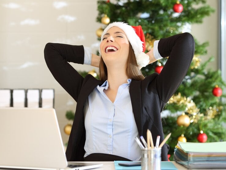 Eine lachende Frau in einem Büro, die eine Weihnachtsmütze trägt, sitzt entspannt zurückgelehnt vor ihrem Laptop. Im Hintergrund steht ein geschmückter Weihnachtsbaum mit goldenen und roten Kugeln.
