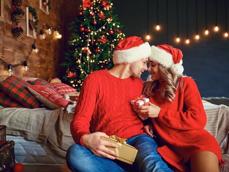 Ein verliebtes Paar in roten Weihnachtsoutfits und Weihnachtsmützen sitzt vor einem festlich geschmückten Weihnachtsbaum. Sie halten sich gegenseitig Geschenke und lächeln sich zärtlich an, während im Hintergrund eine gemütliche, weihnachtliche Szenerie mit Lichterketten und dekorierten Kissen zu sehen ist.