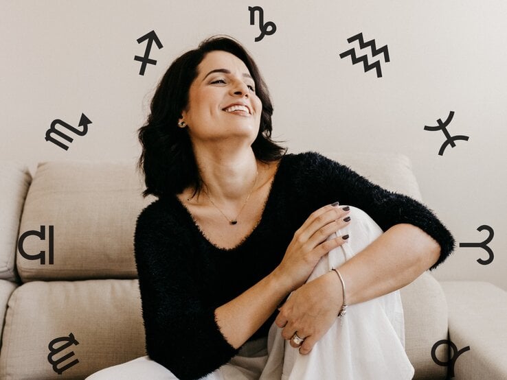 Eine Frau sitzt auf einem Sofa und blickt lachend nach oben. Sie sieht glücklich aus und ist umgeben von den astrologischen Symbolen der Tierkreiszeichen.