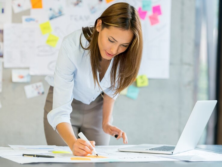 Junge Frau in Business-Kleidung arbeitet konzentriert an einem Schreibtisch, schreibt Notizen auf ein großes Papier und hat einen Laptop sowie bunte Haftnotizen im Hintergrund.