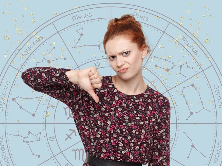 Junge Frau mit roten Haaren und floralem Oberteil, die mit einem unzufriedenen Gesichtsausdruck den Daumen nach unten zeigt. Im Hintergrund ist ein astrologisches Sternzeichenrad mit goldenen Glitzereffekten auf hellblauem Hintergrund zu sehen.