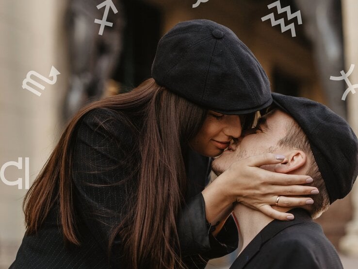Ein verliebtes Paar in dunkler Kleidung und passenden schwarzen Mützen, das sich zärtlich küsst. Die Frau hält das Gesicht des Mannes liebevoll mit beiden Händen. Im Hintergrund sind dezent astrologische Sternzeichensymbole zu sehen.