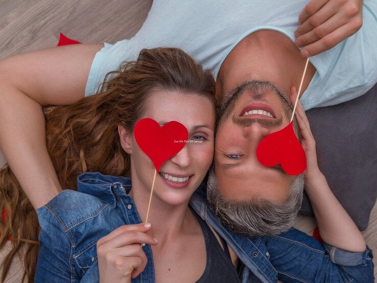Ein verliebtes Paar liegt entspannt auf dem Boden, Kopf an Kopf. Beide lächeln fröhlich und halten jeweils ein rotes Papierherz vor eines ihrer Augen. Die Frau hat lange, wellige braune Haare und trägt ein Jeanshemd, der Mann hat graues Haar, einen leichten Bart und trägt ein hellblaues T-Shirt. Die Stimmung ist verspielt und liebevoll.
