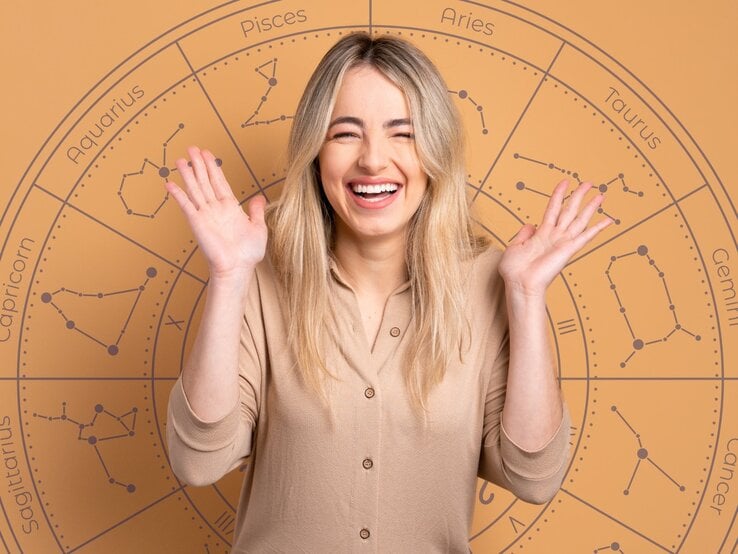 Eine fröhliche, blonde Frau mit langen Haaren lächelt breit und hebt beide Hände freudig in die Luft. Sie trägt ein beigefarbenes Oberteil. Im Hintergrund ist ein großes astrologisches Rad mit den Symbolen und Namen der Sternzeichen wie Aries, Taurus, Gemini und Pisces zu sehen. Die warmen, erdigen Farbtöne des Bildes vermitteln eine positive und optimistische Stimmung.