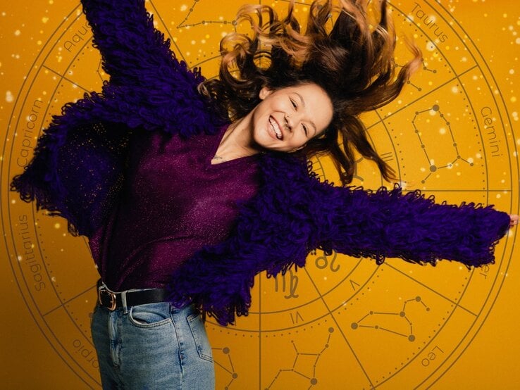 Eine fröhliche Frau mit langen, welligen Haaren springt lächelnd in die Luft. Sie trägt eine violette, flauschige Jacke und ein glitzerndes Oberteil, kombiniert mit einer Jeans. Der Hintergrund ist gelb mit einem astrologischen Tierkreis-Muster, das verschiedene Sternzeichen und Sternbilder zeigt.