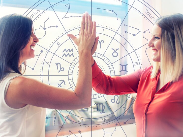  Zwei lächelnde Frauen geben sich ein High-Five vor einem Hintergrund mit einem astrologischen Tierkreis-Muster, das verschiedene Sternzeichen und Symbole zeigt. Die Frau links trägt ein weißes Oberteil, die Frau rechts eine rote Bluse. Die Szene wird von natürlichem Licht durchflutet, das durch Fenster im Hintergrund fällt. Die Geste und die fröhlichen Gesichter vermitteln ein Gefühl von Teamwork und positiver Energie. Der astrologische Hintergrund deutet auf einen Zusammenhang mit Horoskopen oder Sternzeichen hin.