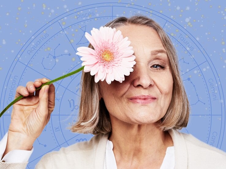 Eine ältere Frau mit schulterlangem, grauem Haar hält eine rosa Blume vor ihr Auge und lächelt sanft. Im Hintergrund ist ein astrologisches Rad mit Sternzeichen und Konstellationen auf einem blauen, sternenübersäten Himmel zu sehen. Das Bild vermittelt eine positive und harmonische Atmosphäre, die Astrologie und Lebensfreude verbindet.