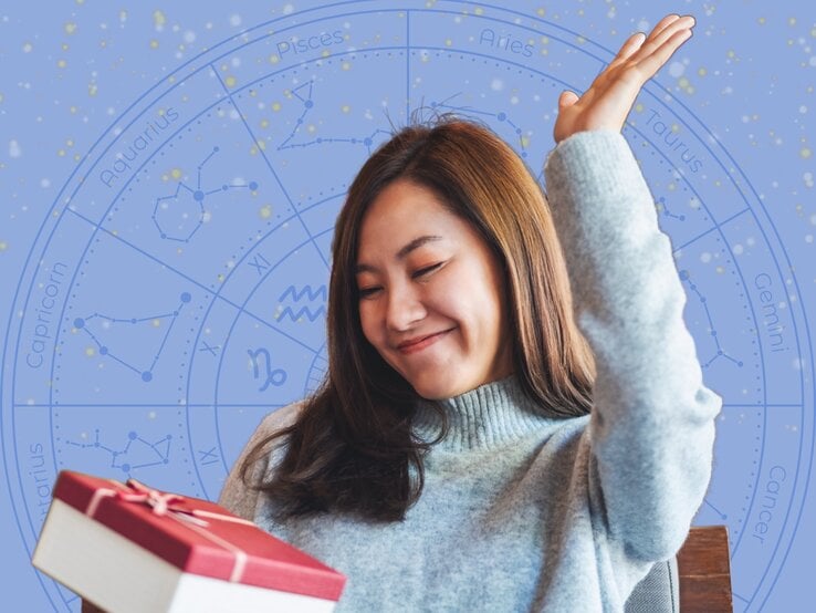 Eine junge Frau mit langem braunem Haar trägt einen hellblauen Strickpullover und hebt freudig eine Hand in die Luft, während sie ein Geschenk mit roter Schleife in der anderen Hand hält. Im Hintergrund ist eine blaue astrologische Sternzeichen-Grafik mit Konstellationen und den Namen der Tierkreiszeichen zu sehen. Die Szene strahlt Glück und Zufriedenheit aus.
