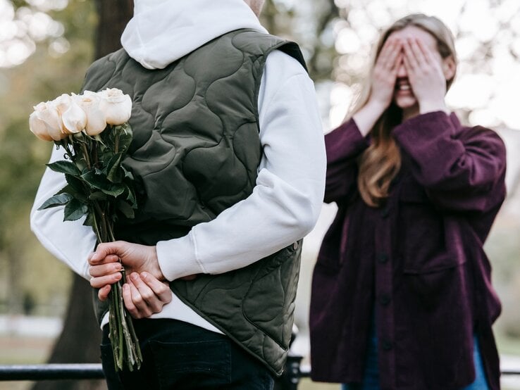 Eine Person in einer weißen Kapuzenjacke und grüner Weste hält hinter ihrem Rücken einen Strauß weißer Rosen, während eine andere Person mit langen Haaren und lila Jacke sich die Augen mit beiden Händen verdeckt und lächelnd auf die Überraschung wartet. Die Szene spielt sich in einem Park mit unscharfem Hintergrund aus Bäumen und Natur ab. Die Atmosphäre wirkt romantisch und verspielt.