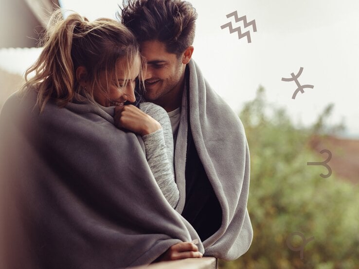 Ein junges Paar steht eng umschlungen auf einem Balkon oder einer Terrasse und teilt eine große, graue Decke. Beide lächeln sich verliebt an, während die Frau schüchtern ihren Kopf senkt. Der Mann lehnt sich sanft zu ihr. Im Hintergrund sind unscharfe grüne Bäume zu sehen, was eine natürliche, gemütliche Atmosphäre schafft. In der oberen rechten Ecke schweben mehrere astrologische Symbole, darunter die Zeichen für Wassermann, Fische und Steinbock, die das Bild mit einer astrologischen Bedeutung versehen.