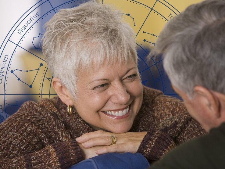 Eine lächelnde ältere Frau mit kurzen, weißen Haaren und goldenen Ohrringen lehnt sich entspannt auf ihre Arme und blickt liebevoll einen Mann an, der ihr gegenüber sitzt. Sie trägt einen warmen, gestrickten Pullover. Der Mann ist leicht unscharf im Vordergrund zu sehen, mit grauem Haar und einem freundlichen Ausdruck. Im Hintergrund befindet sich ein großes astrologisches Rad mit den Tierkreiszeichen und Sternbildern in Blau und Gelb. Das Bild verbindet Romantik und Astrologie, als Symbol für Liebe und Partnerschaft im Zeichen der Sterne.