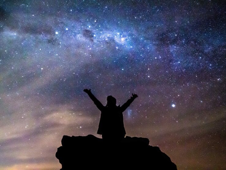 Eine Silhouette einer Person mit ausgestreckten Armen steht auf einem Felsen unter einem beeindruckenden Sternenhimmel. Die Milchstraße leuchtet hell über ihr, während der Himmel in tiefen Blau- und Violetttönen schimmert. Die Szene strahlt ein Gefühl von Freiheit, Staunen und Verbundenheit mit dem Universum aus.