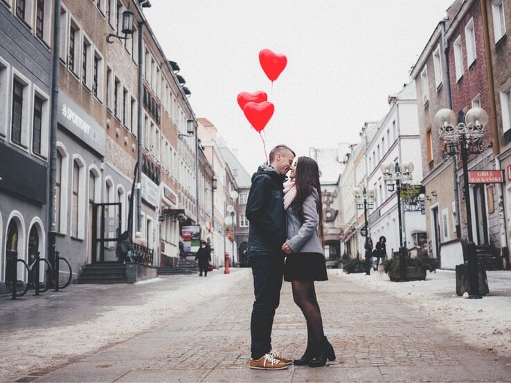 Ein junges Paar steht in einer winterlichen Altstadtstraße, eng umschlungen und hält sich an den Händen. Der Mann trägt eine dunkle Jacke und Sneaker, die Frau einen grauen Mantel, einen Schal und hohe Stiefel. Sie schauen sich verliebt in die Augen. Über ihnen schweben drei rote Herzballons. Die Straße ist gesäumt von historischen Gebäuden, Laternen und einigen Fußgängern im Hintergrund.
