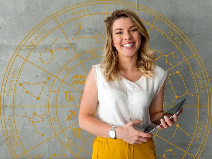 Eine junge Frau mit schulterlangem, welligem blondem Haar steht lächelnd vor einer grauen Wand mit einem goldenen astrologischen Tierkreis. Sie trägt eine weiße Bluse, eine gelbe Hose und eine silberne Uhr am Handgelenk. In ihren Händen hält sie ein Tablet. Der Hintergrund zeigt die Sternzeichen mit ihren Symbolen und Sternbildern in einem kreisförmigen Design.