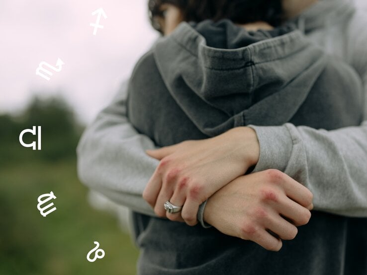 Ein Paar umarmt sich innig im Freien, beide tragen graue Hoodies. Die Hände der Personen sind sichtbar, mit leicht geröteten Knöcheln. Im Hintergrund sind unscharfe Bäume oder Natur zu erkennen. Um das Paar schweben astrologische Symbole verschiedener Sternzeichen, was dem Bild eine mystische, horoskopbezogene Atmosphäre verleiht.