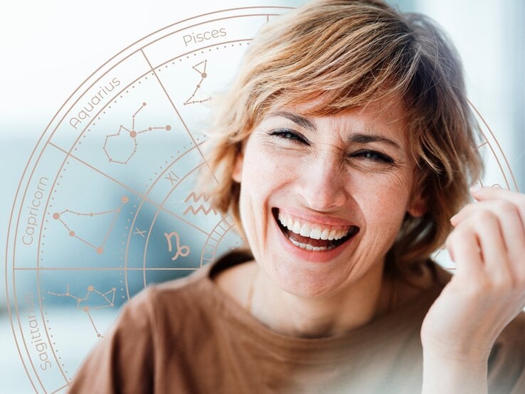 Fröhliche Frau mit kurzen, blonden Haaren lacht herzlich vor einem dezenten astrologischen Sternzeichen-Rad. Der Hintergrund ist hell und freundlich, das Rad mit den Tierkreiszeichen schimmert in sanften Pastelltönen. Das Bild strahlt Optimismus, Lebensfreude und eine positive astrologische Botschaft aus.