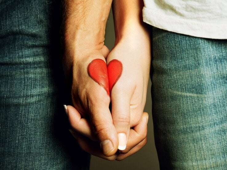 Nahaufnahme von zwei Händen, die sich halten. Auf den Handrücken ist jeweils eine Hälfte eines roten Herzens gemalt, das sich beim Händchenhalten zu einem vollständigen Herz zusammenfügt. Beide Personen tragen Jeans, eine Person trägt ein helles Shirt. Symbolische Darstellung von Liebe und Verbundenheit.