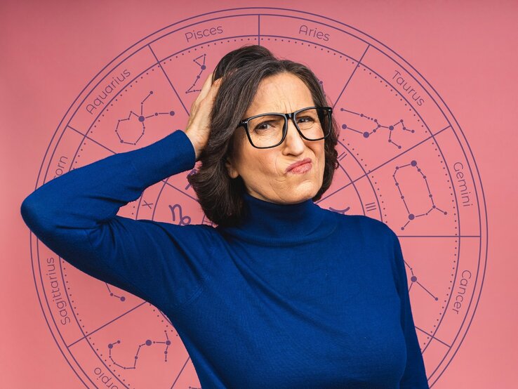 Eine Frau mittleren Alters mit Brille und dunklem Haar trägt einen blauen Rollkragenpullover und hat einen skeptischen, nachdenklichen Gesichtsausdruck. Sie kratzt sich am Kopf, als wäre sie unsicher oder verwirrt. Im Hintergrund befindet sich eine astrologische Tierkreisgrafik mit den Sternzeichen und ihren Symbolen auf einem rosa Hintergrund. Das Bild vermittelt eine humorvolle oder fragende Haltung gegenüber Horoskopen und Astrologie.