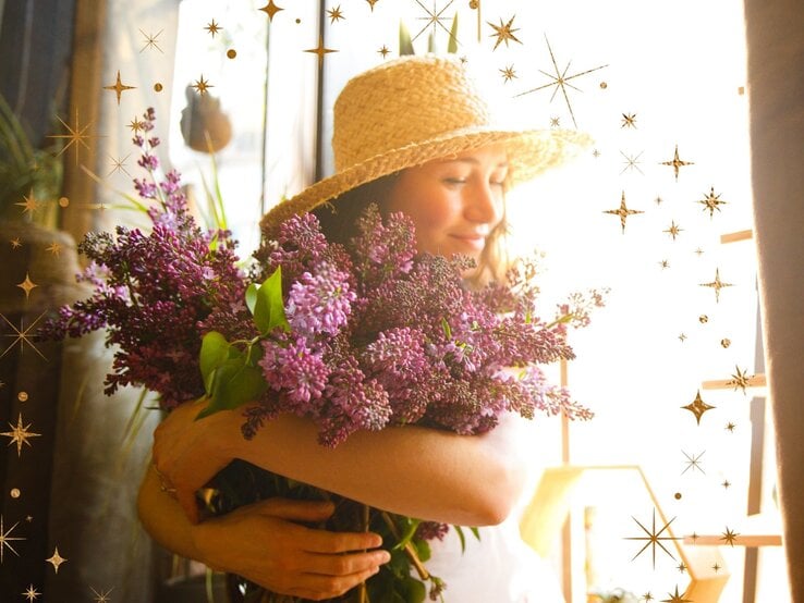 Lächelnde Frau mit Strohhut steht im Sonnenlicht und umarmt einen großen Strauß Fliederblüten. Um sie herum sind goldene, gezeichnete Sterne und funkelnde Elemente eingefügt, die der Szene eine magisch-verträumte Stimmung verleihen.