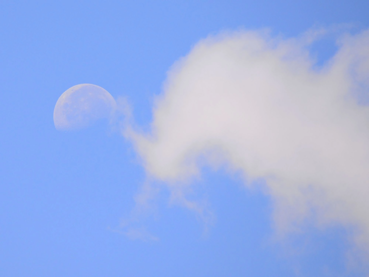Halbmond am hellblauen Himmel, teilweise von einer weißen Wolke verdeckt.