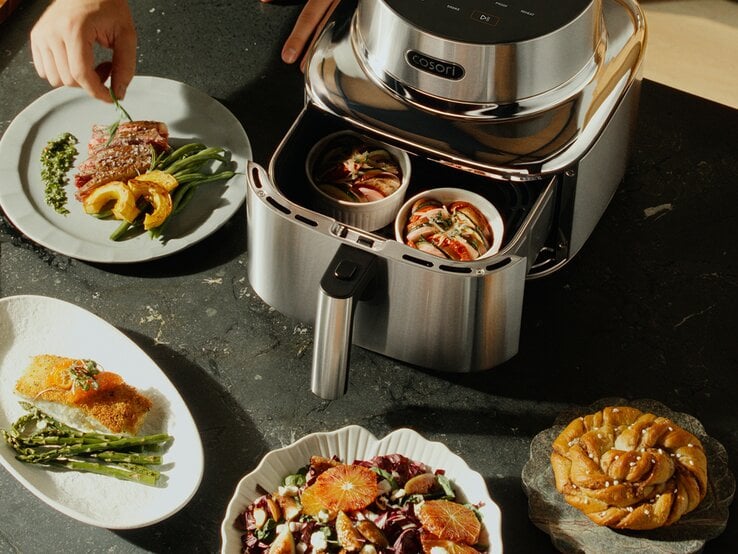 Moderne Heißluftfritteuse auf dunkler Granitfläche, umgeben von angerichteten Gerichten wie Salat, Steak und Gebäck.