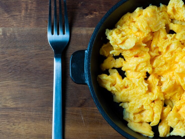 Cremiges Rührei in einer schwarzen Pfanne, angerichtet auf einem Holzbrett mit einer Gabel daneben.