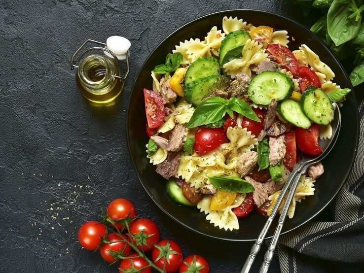 Ein Teller mit Nudelsalat aus Farfalle, Tomaten, Gurken und Thunfisch auf dunklem Untergrund, daneben eine Flasche Olivenöl und Kirschtomaten. | © Getty Images/Lilechka75