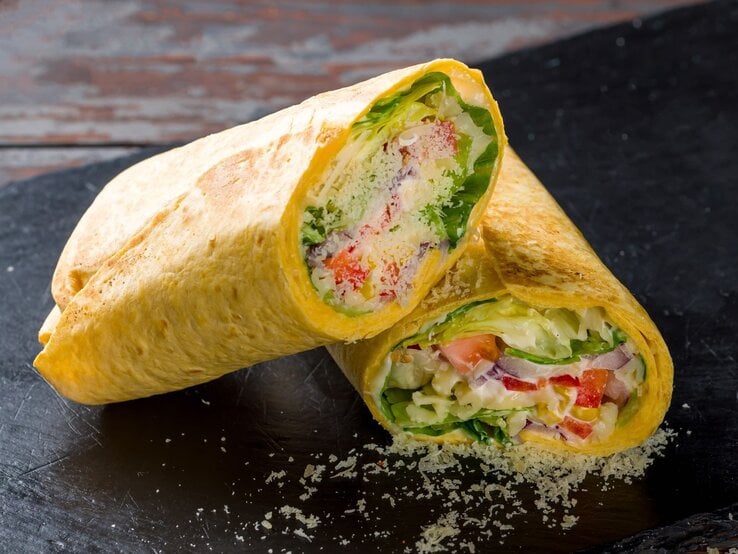 Zwei aufgeschnittene Tortillas gefüllt mit frischem Salat, Tomaten, Zwiebeln und Käse auf einer schwarzen Schieferplatte. | © Shutterstock/SnapFocus