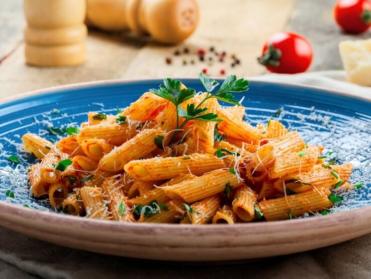Penne in leuchtender Tomatensauce mit Parmesan und Kräutern serviert auf einem blauen Teller vor rustikalem Hintergrund.