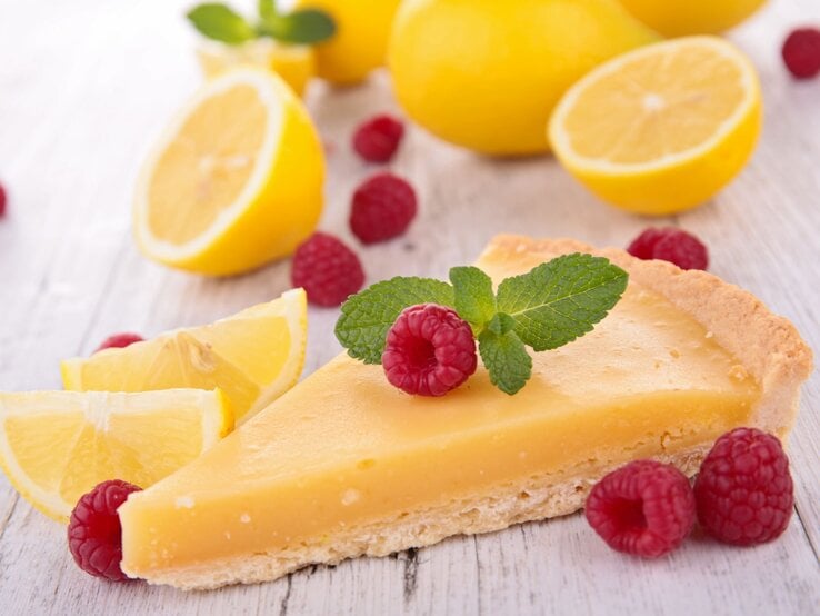 Zitronenkuchen-Stück im Vordergrund, belegt mit Himbeeren und im Hintergrund aufgeschnittene Zitronen