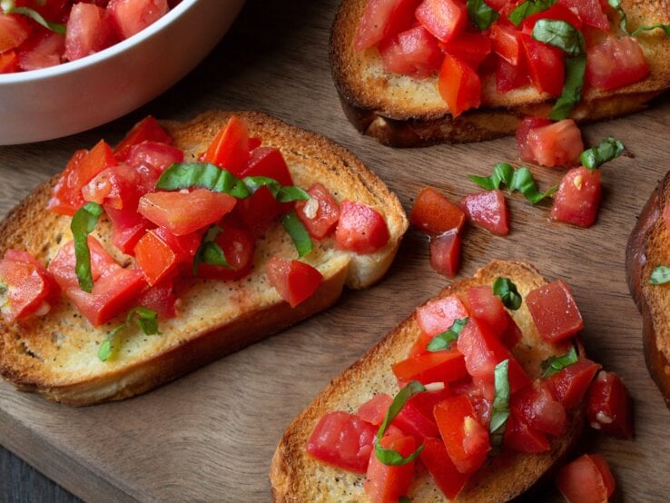 Knusprige Bruschetta mit frischen Tomatenwürfeln und Basilikumblättern auf Holzbrett neben einer weißen Schüssel.