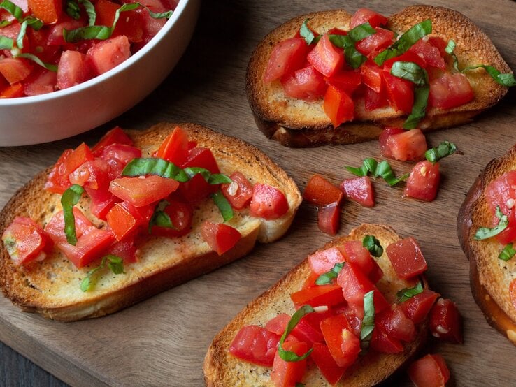 Knusprige Bruschetta mit frischen Tomatenwürfeln und Basilikumblättern auf Holzbrett neben einer weißen Schüssel. | © Adobe Stock/ Catherine Murray