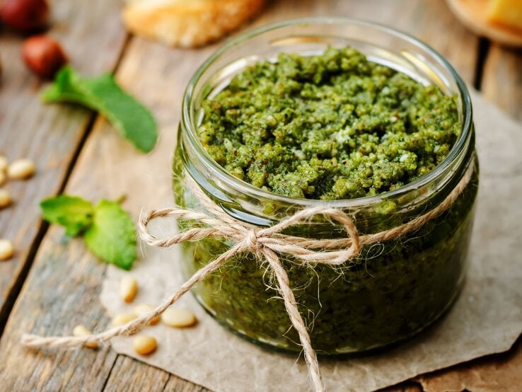 Ein Glas frisches, grünes Pesto mit rustikaler Bastschleife steht auf einem Holzbrett, umgeben von Pinienkernen und Minzblättern.