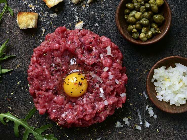 Klassisches Rintertatar / rohes, aufgeschlagenes Ei / Gewürze / schälchen mit fein geschnittenen rohen Zwiebel und kleinen Kapern / Brotwürfel und Ruccolablättern