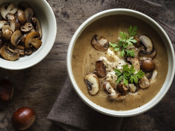 Dunkler Untergrund mit einer Schüssel der Suppe sowie frischen Champignons und Esskastanien drum herum. Alles direkt von oben fotografiert.