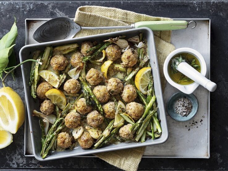 Ein kleines Blech mit dem Spargel und den Hackbällchen auf einem grauen Steintisch. Alles direkt von oben fotografiert.