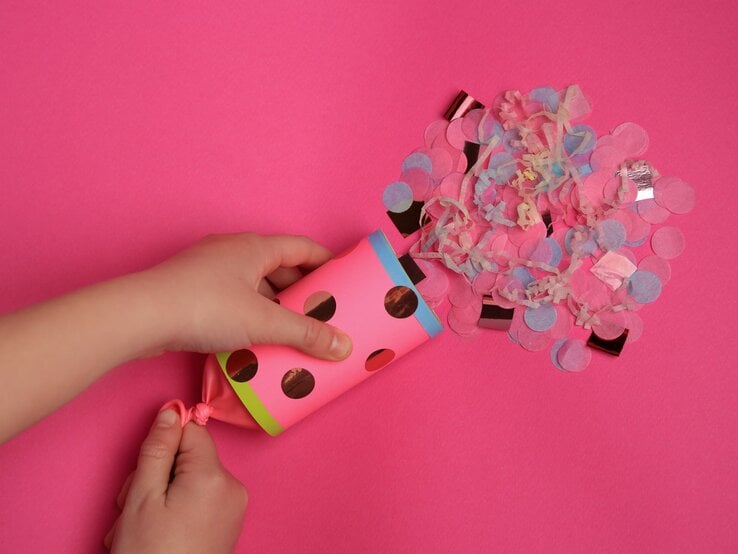 Hände halten eine selbstgebastelte Konfetti-Kanone aus Pappe über einen pinken Hintergrund, aus der gerade buntes Konfetti herausströmt, was eine lebhafte und festliche Atmosphäre erzeugt.