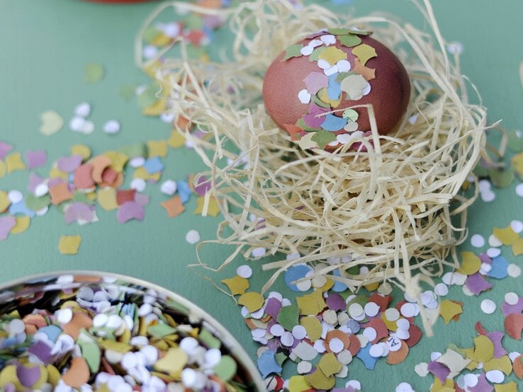 Das Bild zeigt ein sorgfältig dekoriertes Osterei, das in einem Nest aus natürlich aussehendem Stroh platziert ist. Das Ei ist mit bunten Konfettistücken verziert, die auf seiner Oberfläche haften und ihm ein festliches Aussehen verleihen.